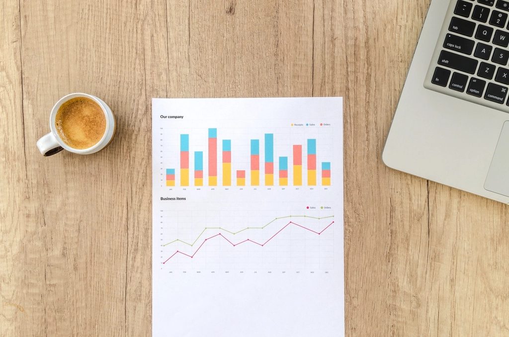 A printed report with colourful bar and line charts on a wooden desk next to a coffee cup and a laptop.