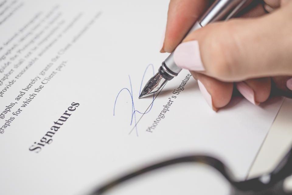 A hand signing the signature line of a printed contract with a silver fountain pen, a pair of glasses blurred in the foreground.