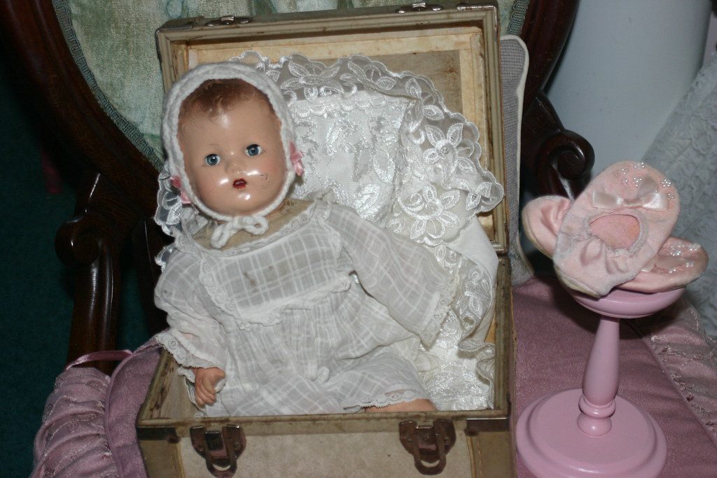 A vintage baby doll in a lace bonnet and gown sitting in an old open trunk, with tiny pink doll shoes on a stand beside it.
