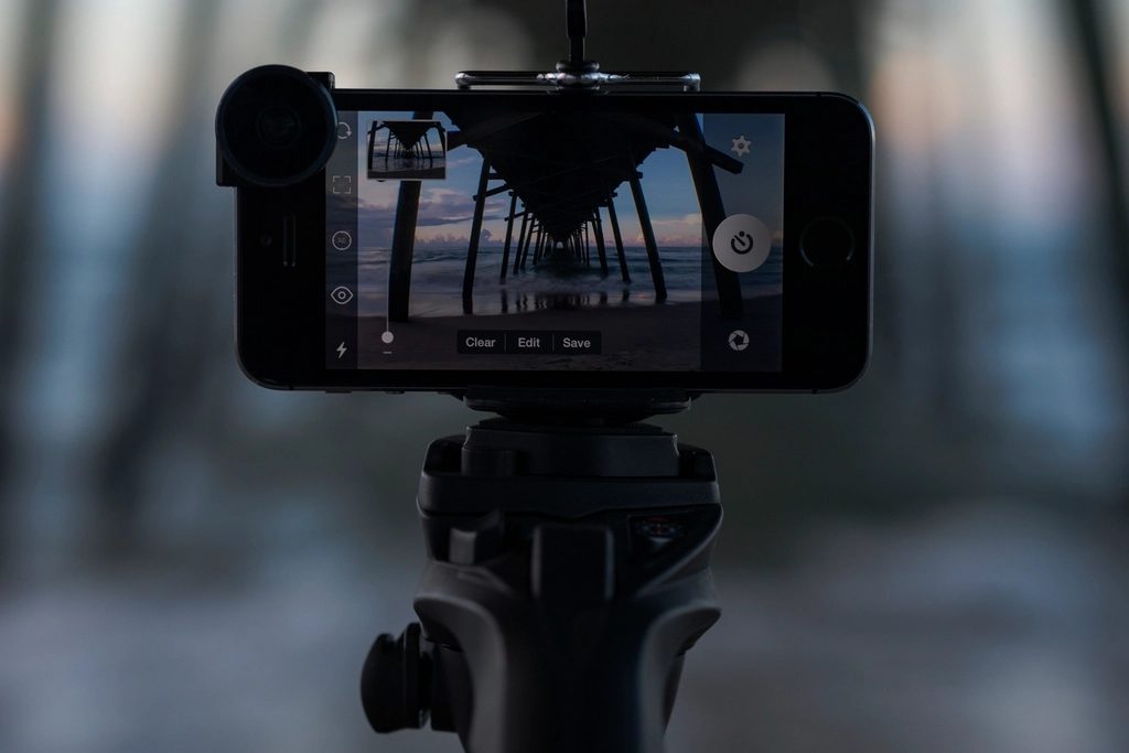 A smartphone clamped sideways on a tripod head, its camera app framing a pier at dusk, with a clip-on lens attached.